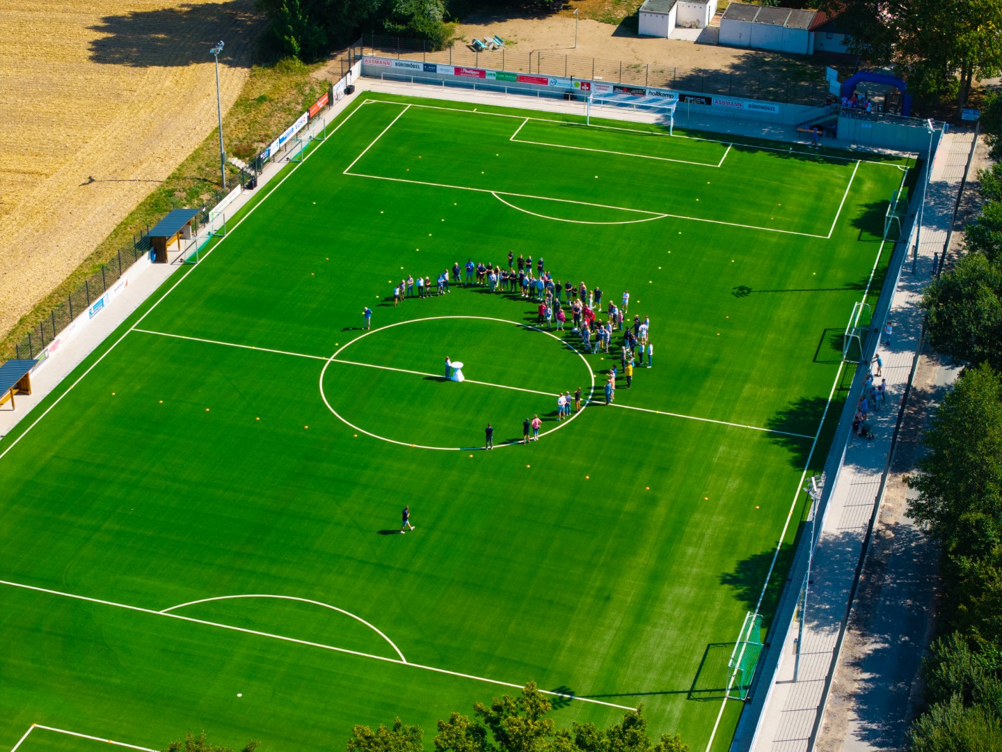 Film über die Einweihung unseres Kunstrasenplatzes am 08.08.2026 Luftaufnahme eines Fußballfelds mit einer Gruppe von Menschen, die in einem Kreis in der Spielfeldmitte stehen