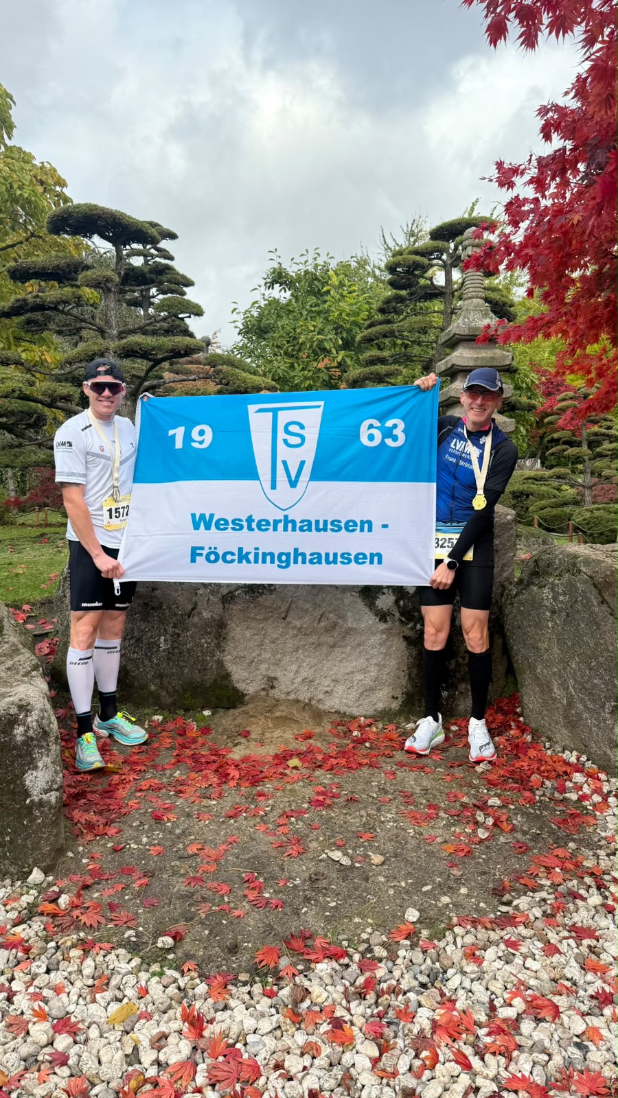 Zwei Männer in Sportkleidung halten eine blau-weiße Fahne mit der Aufschrift 'TSV 1963 Westerhausen-Föckinghausen' vor einer Gartenlandschaft mit roten Blättern am Boden