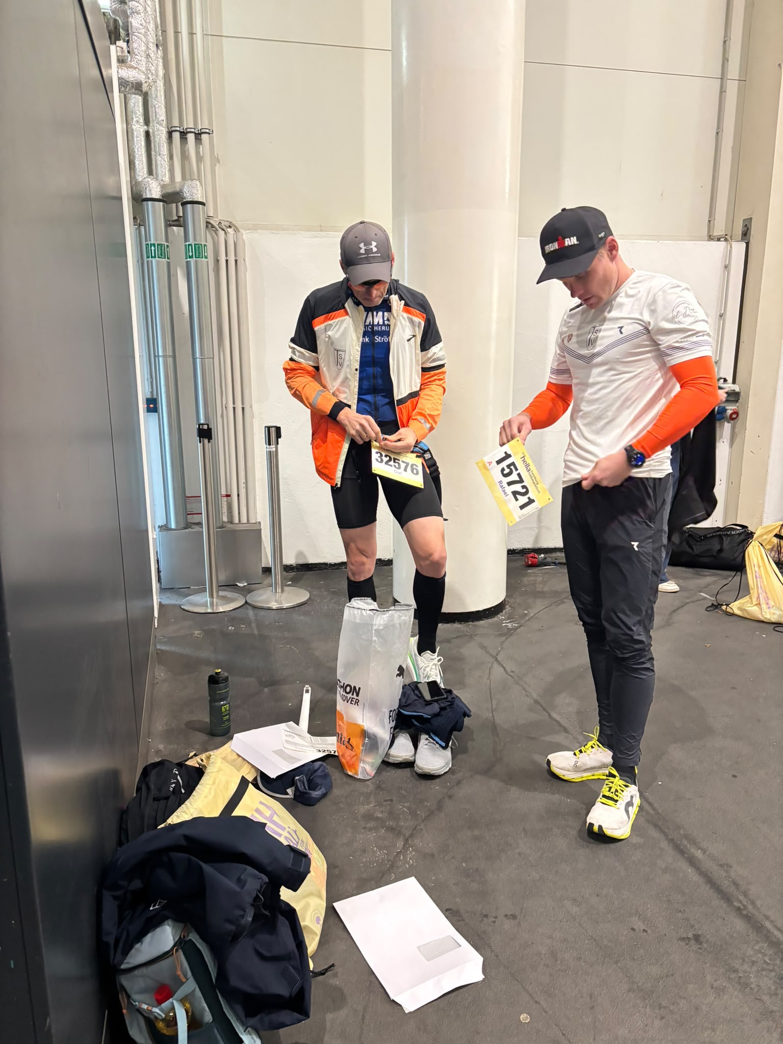 Zwei Sportler in Sportkleidung und Mützen bereiten sich in einer Halle vor, einer hält eine Startnummer in der Hand