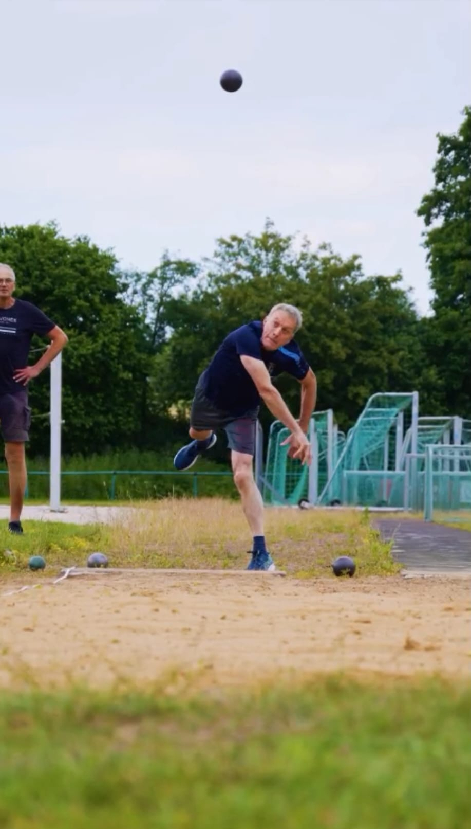 Sportabzeichen beim TSV Mann wirft Kugel beim Kugelstoßen auf einer Sportanlage mit Gras und Bäumen im Hintergrund